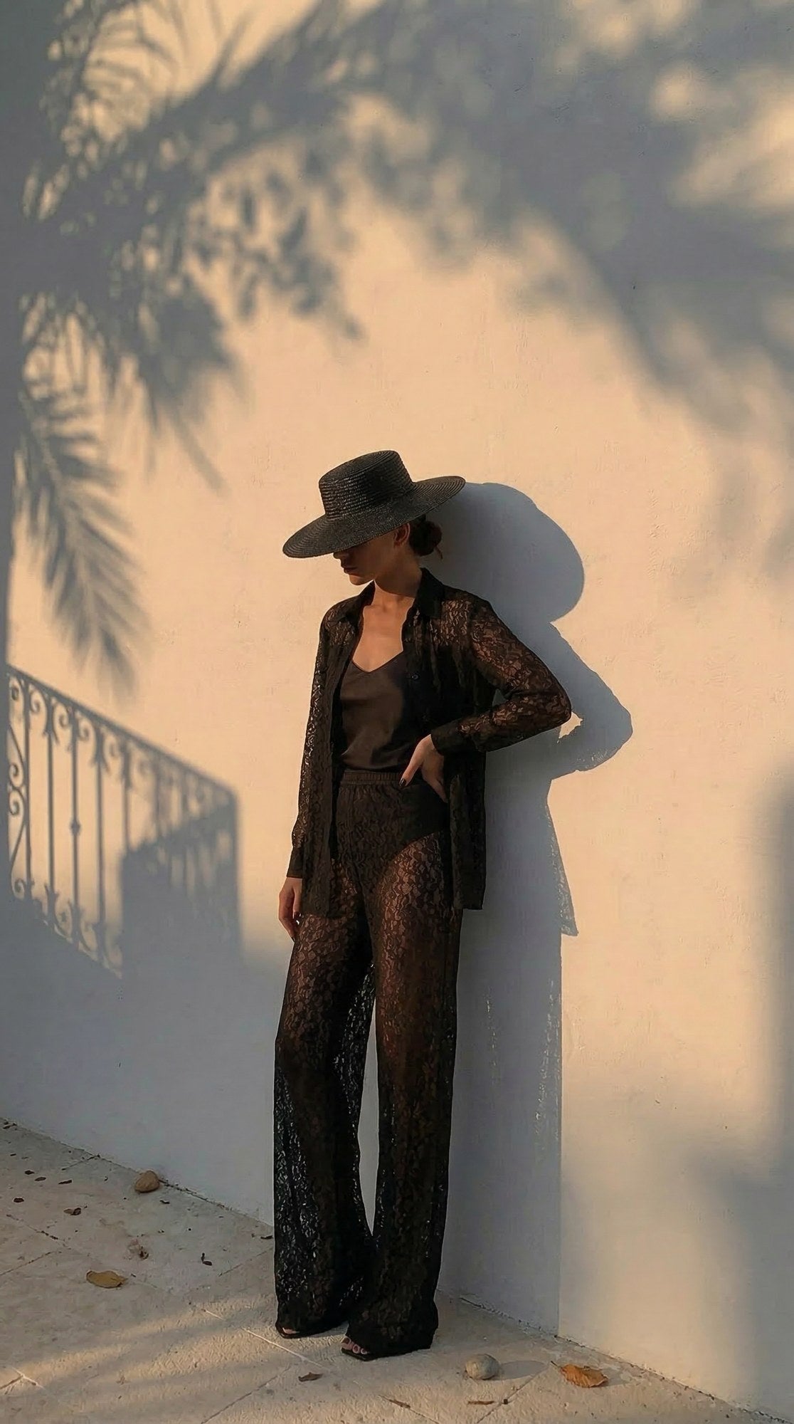 Model in the Lace Set shirt and trousers against a sunlit wall with a wide-brim hat